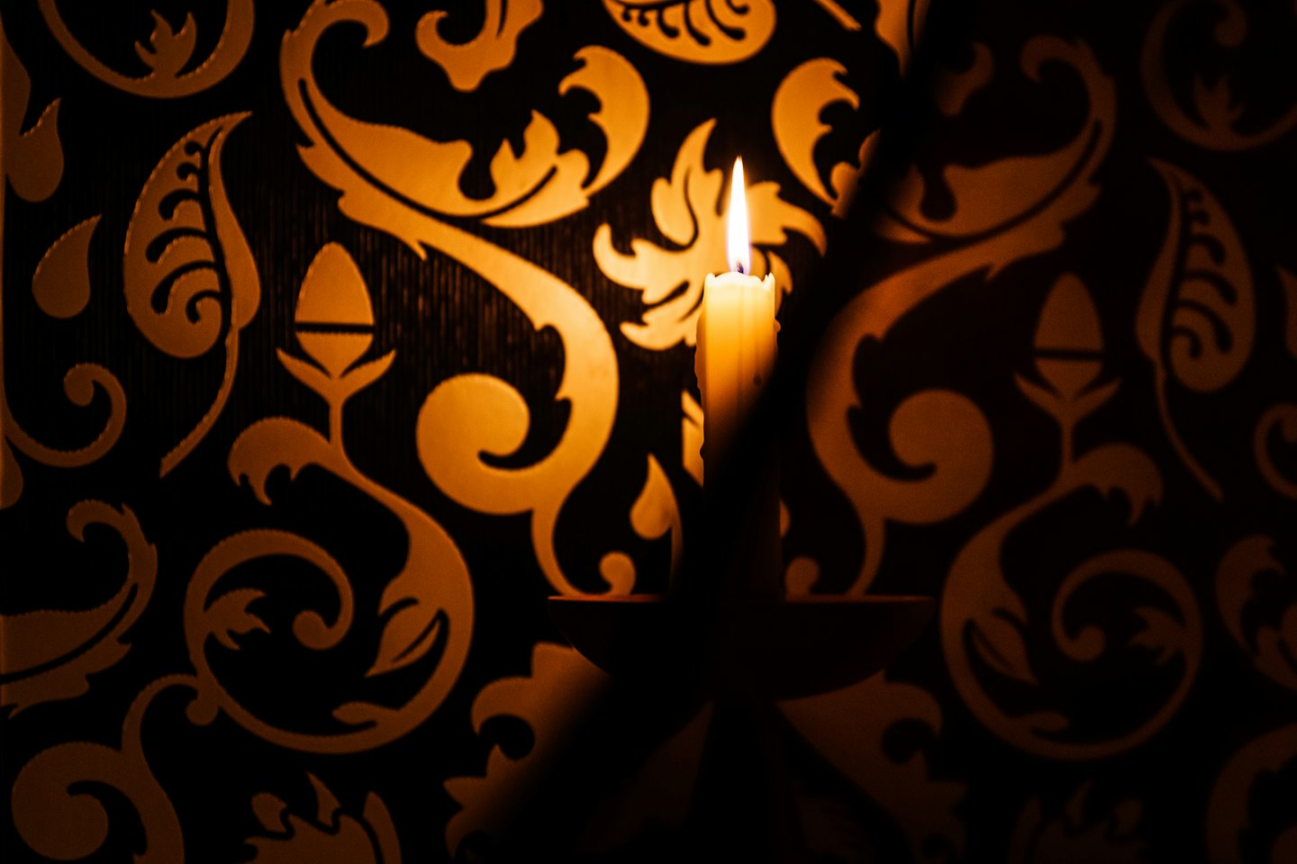 A lit white candle sits against patterned floral wallpaper.