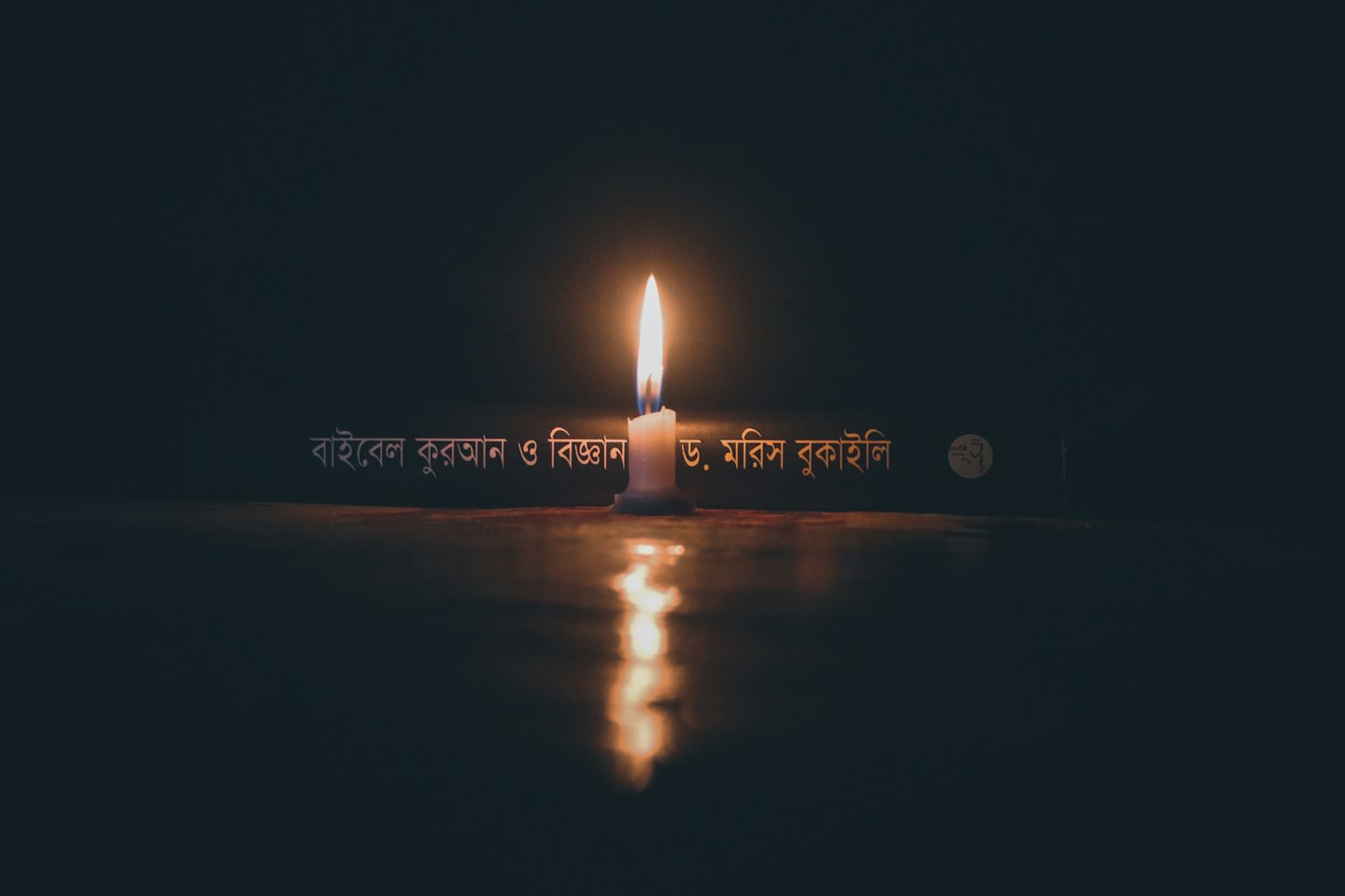 A white candle glows on a dark wooden table near a book.